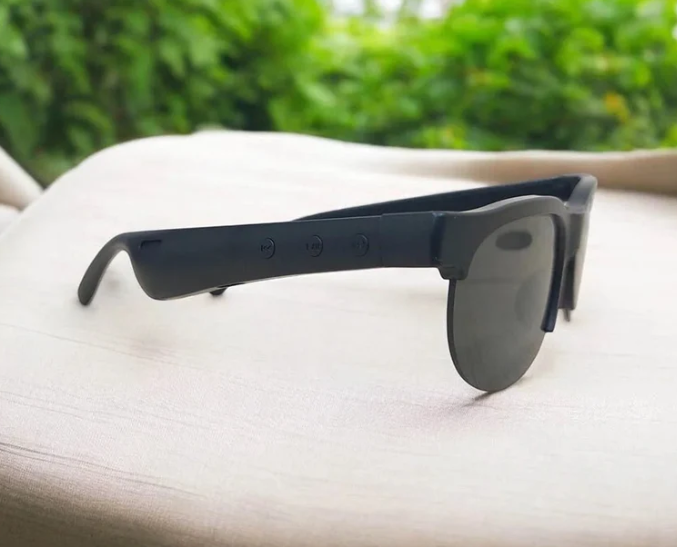 Black smart sunglasses with a matte finish and dark lenses, resting on a light beige fabric surface with a blurred green outdoor background.